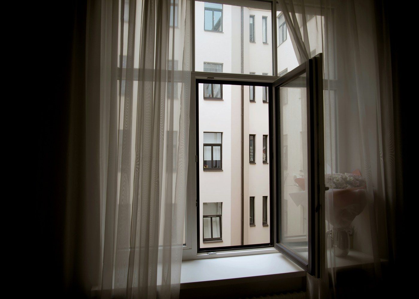 An open window with sheer curtains looks onto a building facade at close range.
